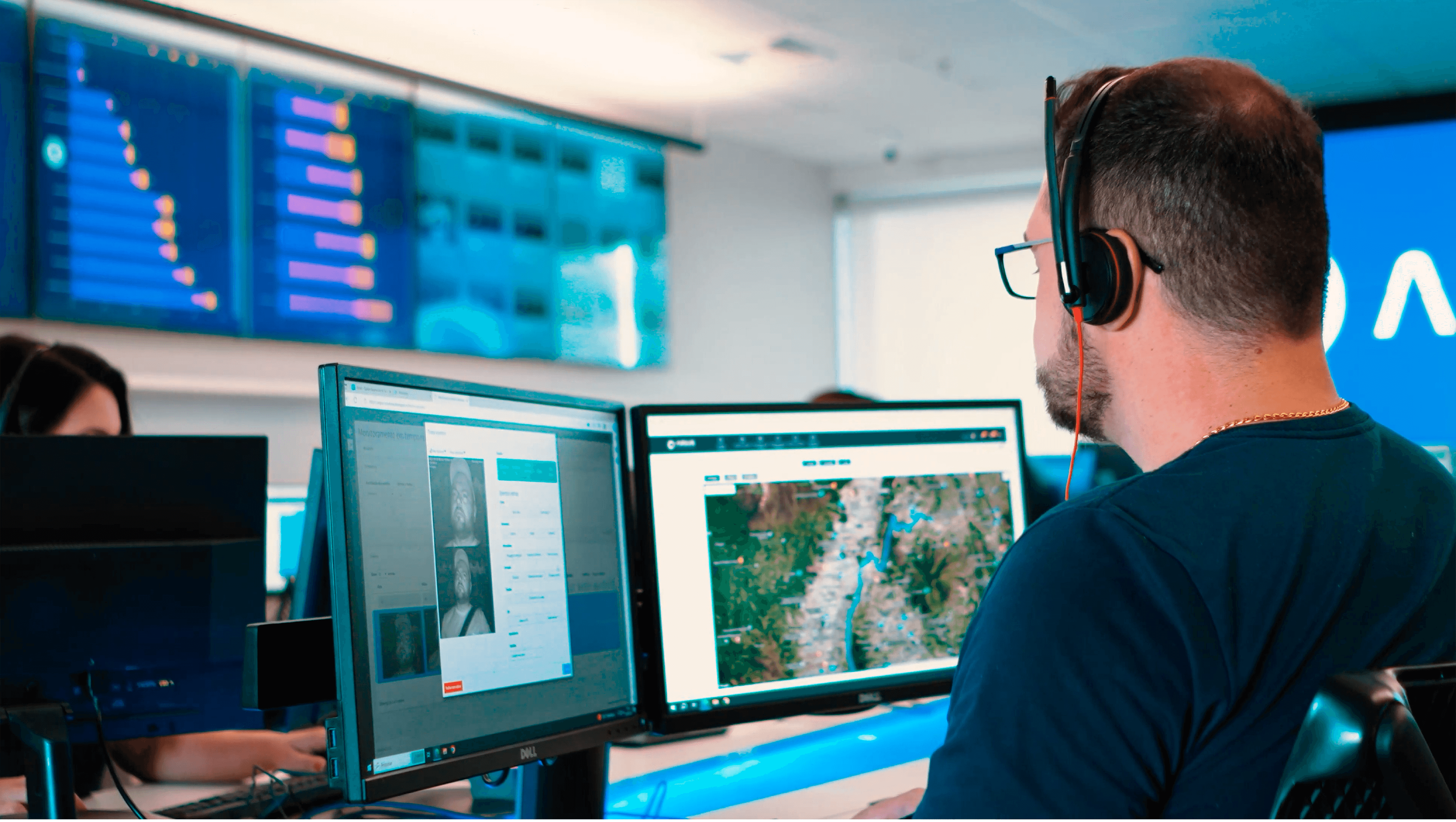 A imagem contém um homem utilizando um computador com dois monitores, no monitor esquerdo está a plataforma da argus e no direito tem um mapa com pontos azuis. O homem está de costas, vestindo uma camisa azul escuro e um headset preto